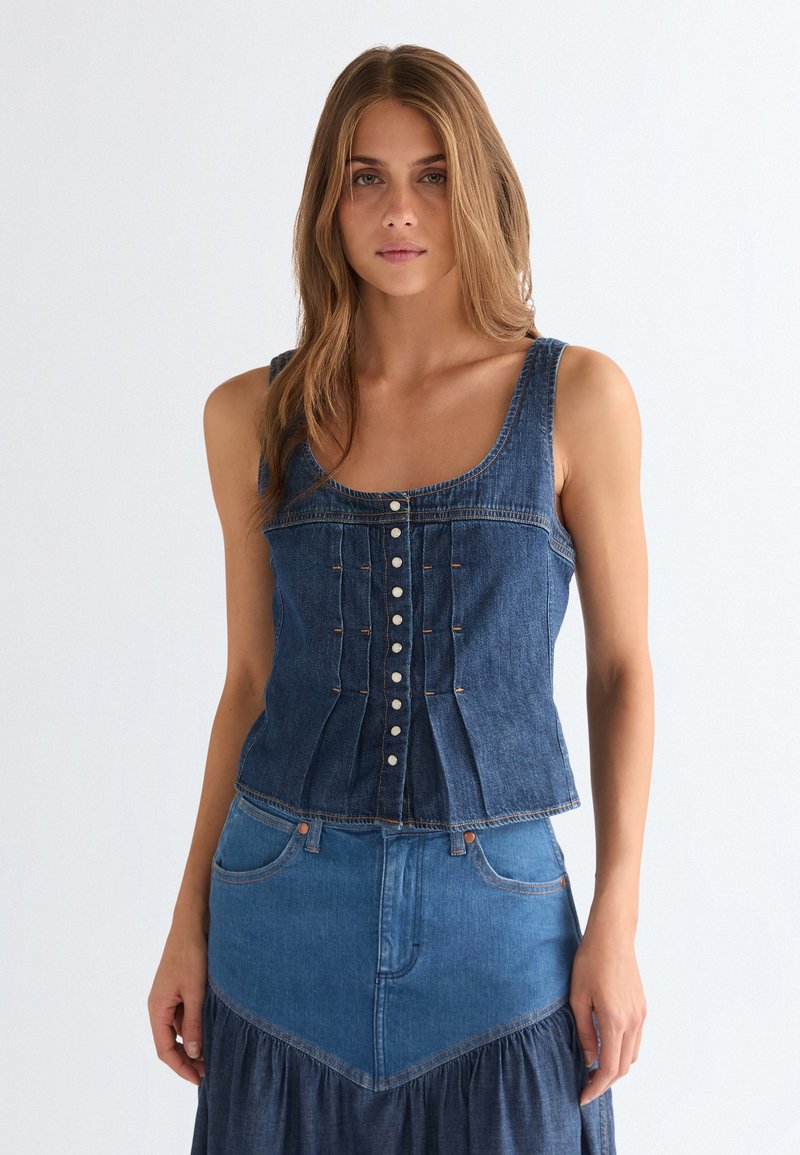 Νέα γυναίκα με μακριά μαλλιά που φοράει σκούρο μπλε τζιν αμάνικο τοπ με κουμπιά και αντιστοιχισμένη τζιν φούστα, κοιτάζοντας μπροστά σε λευκό φόντο.