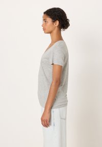 Mlada ženska s kodrastimi lasmi, oblečena v sivo kratkoročno majico z V-izrezom in svetlomodre kavbojke, stoji v profilu pred preprostim belim ozadjem.