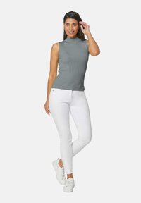 Mouwloze grijze ribbeltrui met een opstaande kraag, gecombineerd met witte skinny jeans en witte sneakers. Model poseert met één hand op haar hoofd.