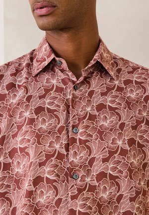 Mann trägt ein braunes Hemd mit hellrosa Blumenmuster, zugeknöpft, Kragen offen, Lippen und Kieferlinie sichtbar.