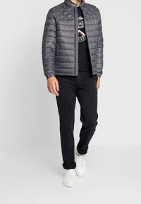Giacca trapuntata grigia con colletto alto e chiusura a zip, abbinata a una camicia nera, pantaloni neri e sneakers bianche.