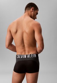 Zwarte heren boxershorts met een brede witte 'CALVIN KLEIN' tailleband, met een gladde textuur en een aansluitend ontwerp, gezien vanuit de achterkant.