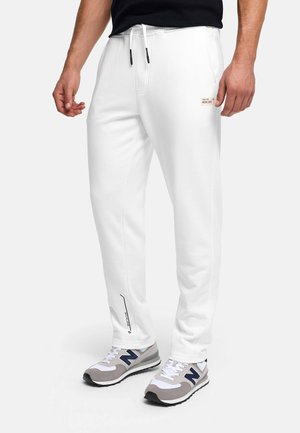 Mężczyzna w białych spodniach dresowych ze ściągaczem i bocznym logo oraz szarych butach New Balance, stojący na białym tle.