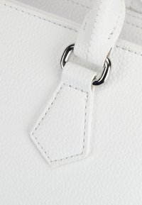 Biała torebka z teksturowanej skóry z bliska prezentuje gładki uchwyt, srebrne okucia oraz sześciokątną skórzaną metkę ze stębnowanymi brzegami.