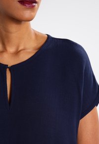 Top bleu marine a maniche corte realizzato in tessuto testurizzato, caratterizzato da un design a occhiello sul collo con dettagli metallici sottili.
