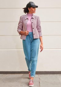 Kariert rosa-weiße Jacke, hellrosa Rollkragenpullover, hellblaue Jeans und rosa Turnschuhe; Außenaufnahme mit neutralem Hintergrund.