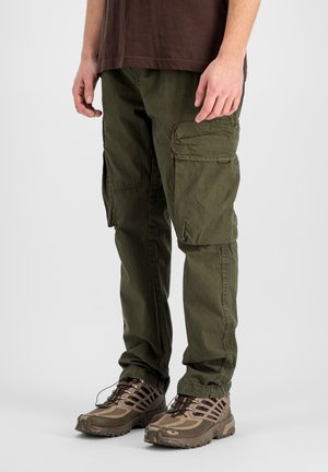 Man wearing olive green cargo pants and brown hiking shoes, standing against a plain light background.