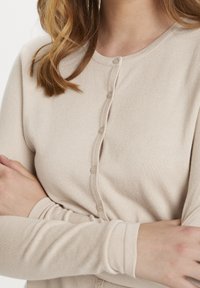 Beige kofta av lätt, ribbad tyg. Har rund halsringning, långa ärmar och en rad små knappar framtill.