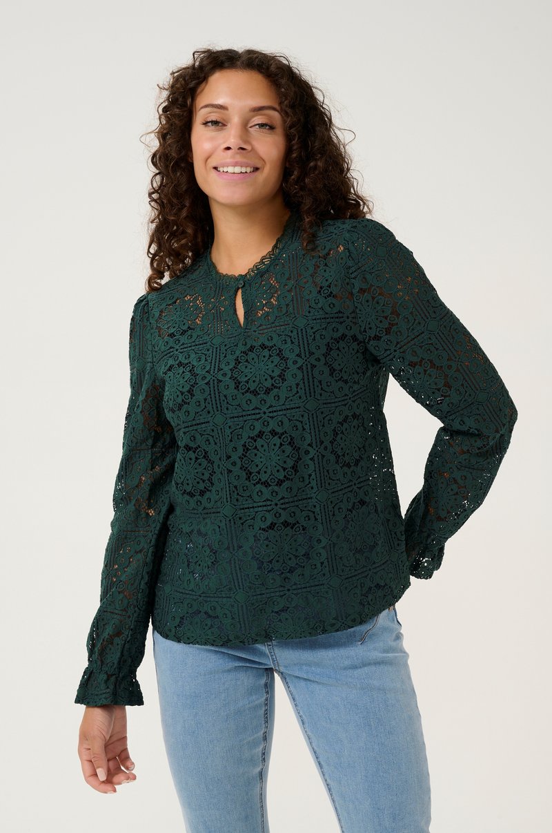 Green lace blouse with intricately detailed floral patterns, keyhole neckline, long sleeves, and a relaxed fit. Paired with blue jeans.