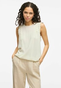 Blusa sin mangas de un suave color beige claro con detalles de encaje en las costuras de los hombros, que presenta una textura suave y un corte holgado, combinada con pantalones beige claros.