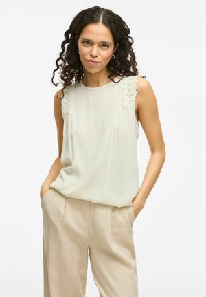 Blouse sans manches couleur beige clair avec des détails en dentelle sur les coutures des épaules, offrant une texture douce et une coupe ample, associée à un pantalon beige clair.