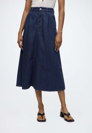 Femme portant une jupe midi en denim bleu foncé à taille haute, associée à des sandales à talons noires ouvertes aux orteils et un haut blanc.