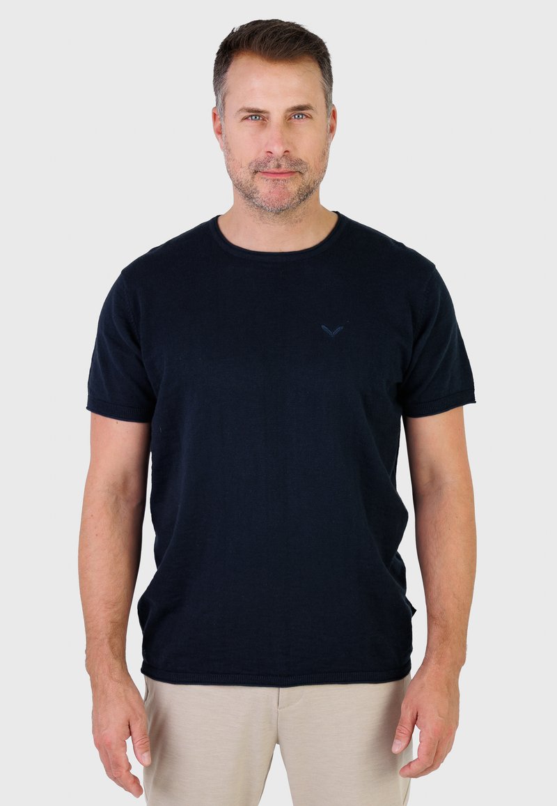 Homme aux cheveux courts et légère barbe de quelques jours, portant un t-shirt noir uni à manches courtes et un pantalon beige, debout devant un fond clair.