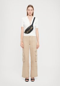 Crni fanny pack s natpisom "LOVE MOSCHINO" u zlatnom textu, nošen preko tijela, u kombinaciji s bijelom rebrastom majicom i bež cargo hlačama. Ravne crne sandale.