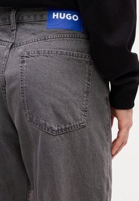 Gray denim jeans with a patch pocket on the back, featuring a blue label with "HUGO" text. Fabric shows a textured finish.