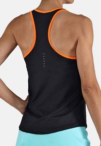 Mujer de espaldas con una camiseta sin mangas negra con ribetes naranjas y pantalones cortos azul claro, con la mano descansando en la cadera.