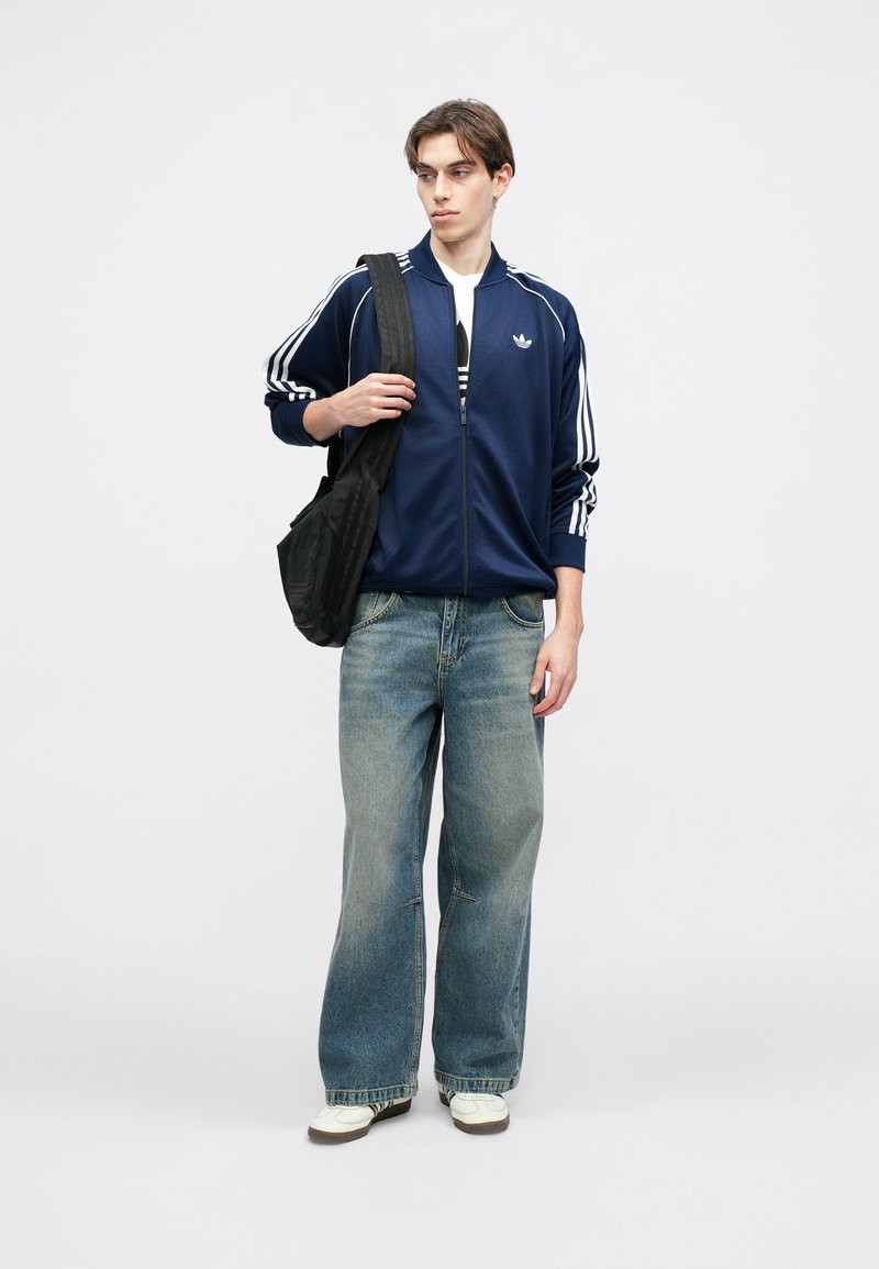 Veste zippée marine avec des rayures blanches, assortie à un jean bleu ample et des chaussures blanches. Le modèle porte un sac noir sur une épaule.