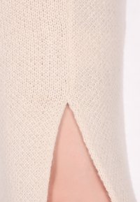 Helles beige Strickgewebe mit einem seitlichen Schlitz, der einen Hauch von Haut zeigt. Die Textur ist weich mit einer feinen, lockeren Webart.