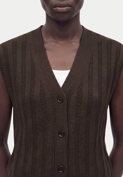 Gilet en tricot côtelé marron foncé avec trois boutons porté sur un top blanc côtelé, vu des épaules jusqu'au milieu du torse.
