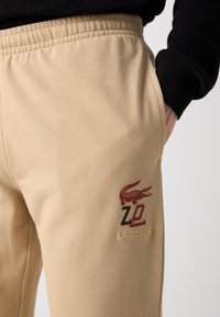 Béžové, textúrované tepláky s elastickým pásom. Obsahujú vyšívané logo Lacoste v červenej a zlatej farbe a štýlizované 'Z' na vrecku.