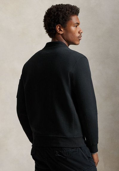 Noor mees lokkis juustega, kellel on seljas must jakk ja mustad laadungpüksid, vaadates küljepealt lihtsal beežil taustal.