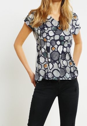 Blouse à manches courtes et décolleté en V en tissu transparent avec un motif circulaire dans des tons marine, gris et terre. Assortie avec un jean noir.