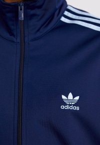 Marinblå zip-up jacka i polyester, med vita tre-rands detaljer på axlarna och en broderad Adidas-logga på bröstet.