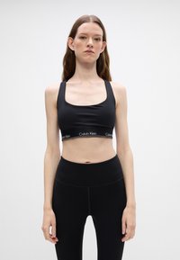 Melns sporta krūšturis, kas izgatavots no gluda materiāla ar platu joslu, kurā ir "Calvin Klein" logotips. Tam ir kvadrāta veida dekoltē un biezas siksnas.