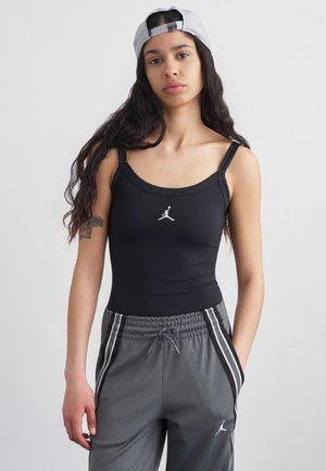 Fiatal nő hosszú sötét hajjal, fehér hátrafordított sapkával, fekete logós ujjatlan pólóval és szürke, oldalsávos szabadidőnadrággal.