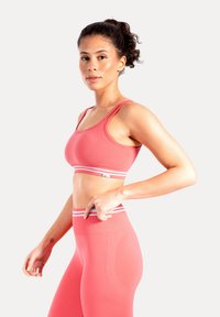 Różowy sportowy biustonosz z białym paskiem w paski; bezszwowy design, dopasowany fason. W komplecie z pasującymi legginsami z wysokim stanem o gładkiej teksturze.