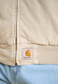 Beige vászon dzseki elülső cipzárral, bordázott derékkal és narancssárga Carhartt logóval. Világoskék farmernadrággal párosítva.