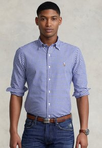 Blue and white checked button-up shirt with rolled sleeves, cotton material, worn with denim jeans and a brown belt and watch.