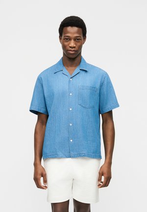 Homme portant une chemise à manches courtes bleu clair à boutons et un short blanc, debout devant un fond clair uni.