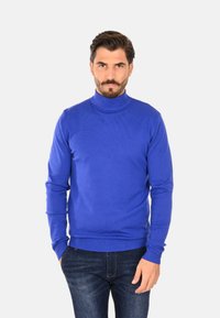 Maglione a collo alto in maglia blu con polsini e orlo a coste, caratterizzato da una texture liscia e un design aderente, indossato con jeans in denim scuro.