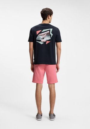 Man draagt een zwart T-shirt met een geometrische windsurfingafbeelding en roze korte broek, staand en met de rug naar de camera op een effen achtergrond.