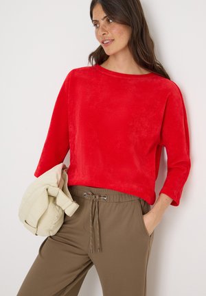 Haut rouge texturé à manches longues avec un décolleté bateau, associé à un pantalon beige à cordon. Un sac couleur crème est tenu dans le bras droit du mannequin.