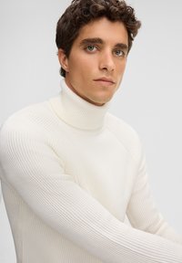 Maglione bianco a collo alto con coste, realizzato in materiale spesso a maglia, caratterizzato da un alto colletto e maniche aderenti, indossato da un modello con capelli ricci corti.