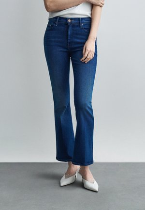 Nő sötétkék bootcut farmerben, fehér felsőben és fehér hegyes orrú magas sarkú cipőben, szürke háttér előtt áll.