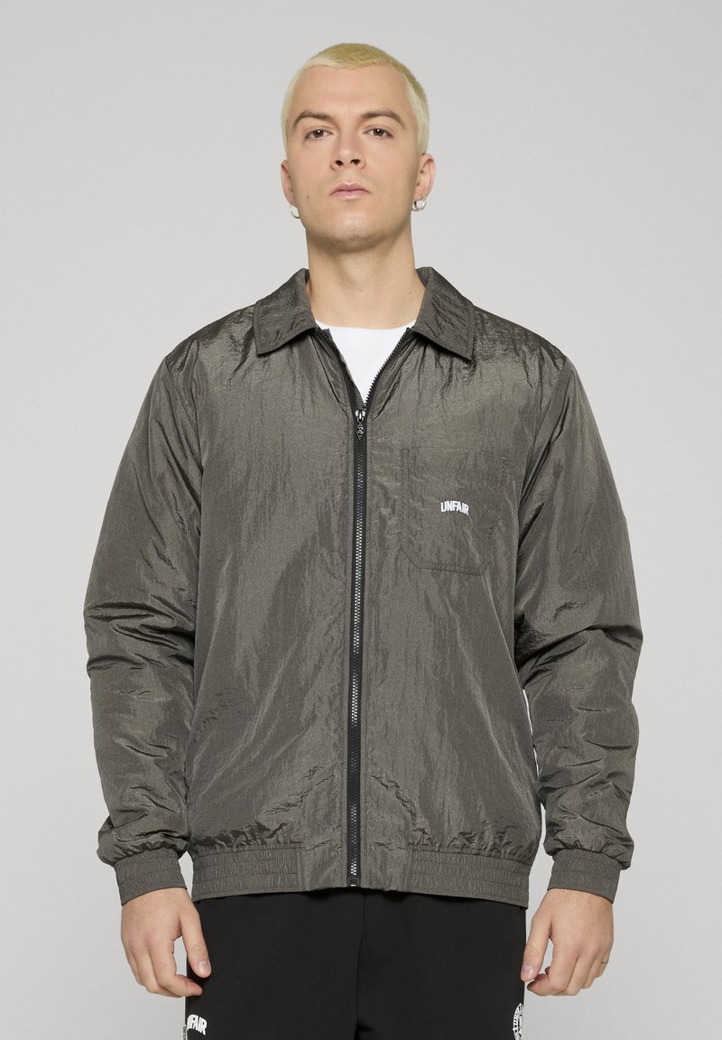 Young person with short blonde hair wearing a shiny dark gray zip-up jacket with "UNFAIR" on the chest pocket.