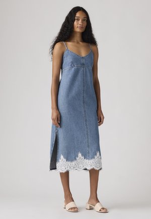 Femme grande aux longs cheveux bouclés portant une robe midi en denim bleu sans manches avec une bordure en dentelle blanche et une fente latérale, associée à des mules à talons blocs beiges.