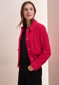 Luddig rosa jacka med krage, framknappar och sidofickor. Buren över en svart topp med strukturerad tyg och avslappnad passform.