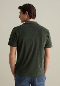 Donkergroen poloshirt met korte mouwen van een gestructureerde stof. Heeft een klassieke kraag en een relaxte pasvorm, gedragen met gestreepte blauw-witte broek.