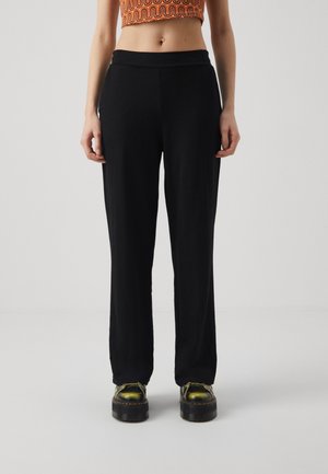 Vrouw met zwarte rechte broek, groene plateau veterschoenen en een cropped oranje top met patroon, staand tegen een effen achtergrond.