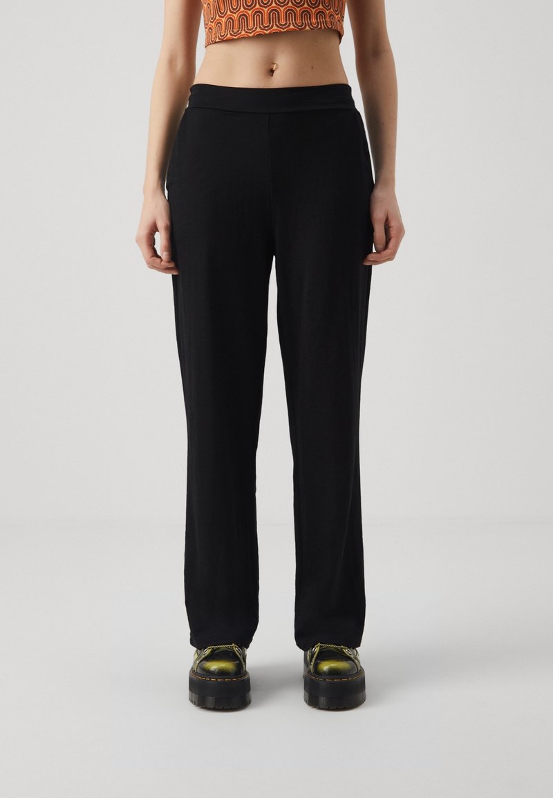 Femme portant un pantalon noir droit, des chaussures à plateforme vertes lacées et un top court orange à motifs, debout devant un fond uni.