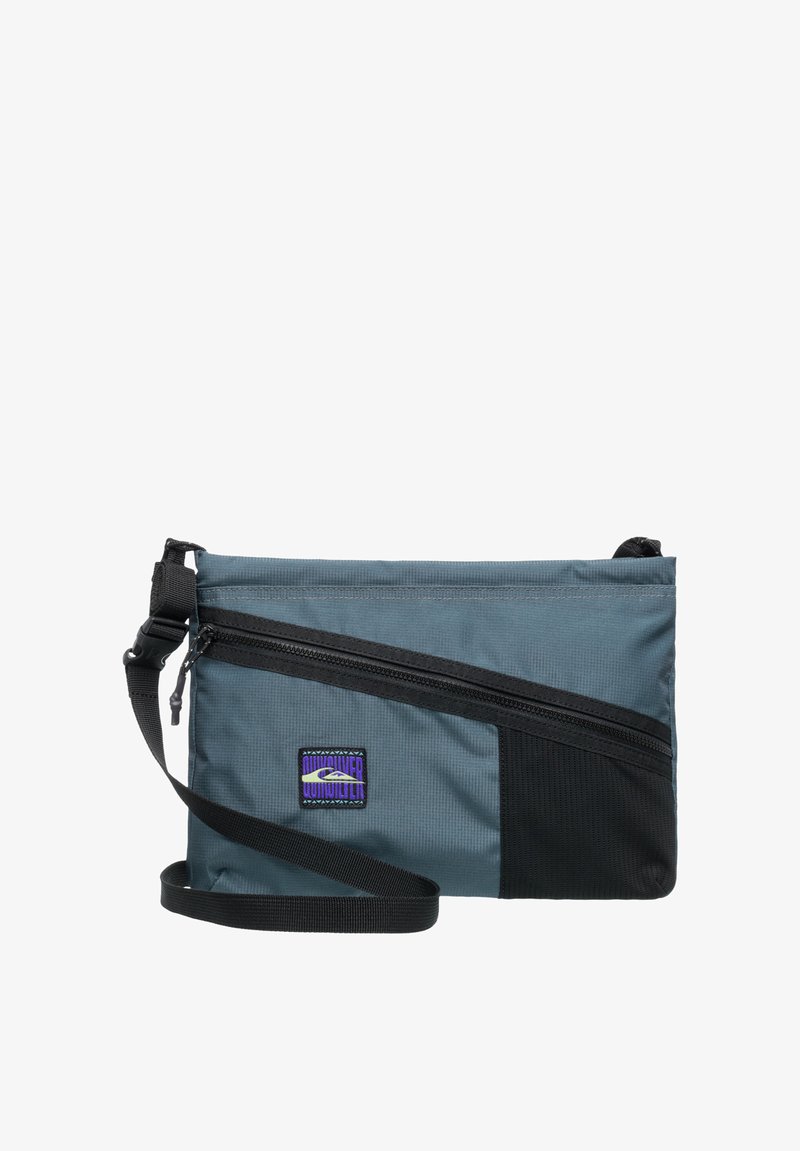 Rektangulär grå tygaxelväska med svarta detaljer, justerbar rem och framficka med dragkedja. Har en logopatch i lila och svart.