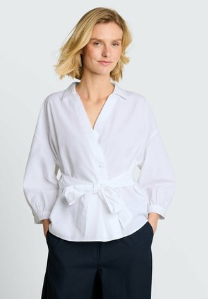 Frau mit kurzen blonden Haaren, die eine weiße Wickelbluse mit einer gebundenen Schleife an der Taille und dunkle Hosen trägt, vor einem schlichten Hintergrund.