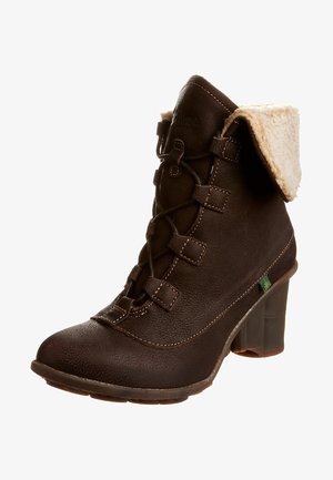 Bota de cuero marrón hasta el tobillo con cuello de piel sintética color beige, cordones en la parte frontal, superficie texturizada y tacón grueso. Detalle de logotipo verde cerca de la suela.