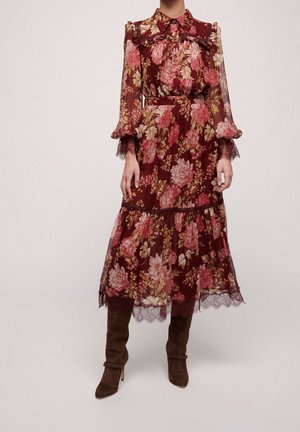 Kobieta nosząca bordową, kwiatową, midi sukienkę z długim rękawem, wykończoną koronką, z kołnierzykiem zapinanym na guziki oraz brązowymi zamszowymi kozaczkami za kolano.