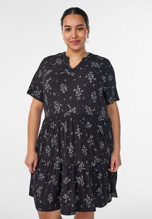 Vrouw met donker haar dat naar achteren is gebonden, draagt een zwarte jurk met korte mouwen en een wit bloempatroon, staand tegen een witte achtergrond.