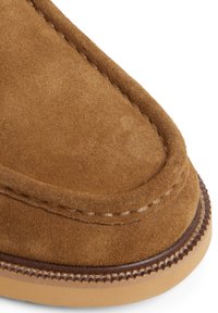Pantoufle en suede marron avec des détails cousus le long du bord et une semelle en caoutchouc beige. Texture lisse avec un bout arrondi et sans fermeture.